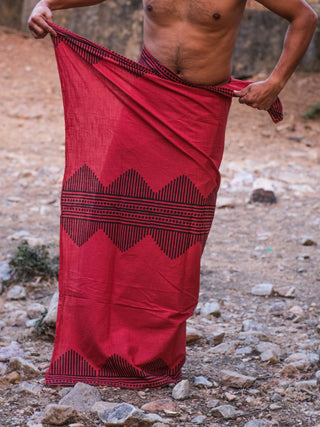 Wearing the Arya Dhoti with a red block print, a person stands outdoors on rocky ground, their upper body bare and face unseen.