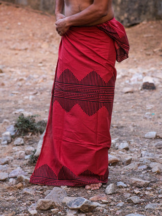 Barefoot on rocky ground, a person wears the Arya Dhoti, styled traditionally with a red and black zigzag block print wrapped at the waist; their upper body is not visible.