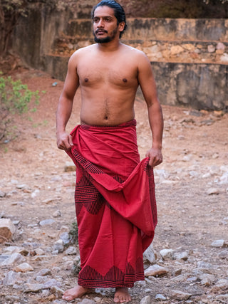 A barefoot, bearded man stands outdoors on rocky ground, wearing a red Arya Dhoti featuring a traditional block print at the waist and no shirt, looking to the side.