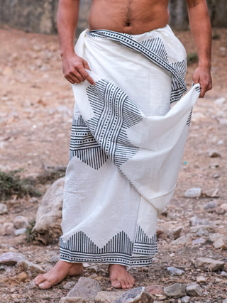 A barefoot person stands outdoors on rocky ground, wearing an Arya Dhoti—a white sarong with a black block print—styled traditionally and holding part of the Arya Dhoti in one hand, their torso bare.