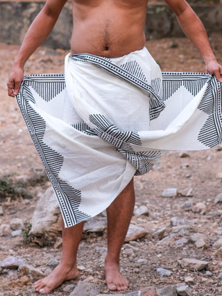A person stands barefoot outdoors, wearing the Arya Dhoti with a black geometric block print draped around their waist, upper body bare, against a backdrop of rocks and dry ground.