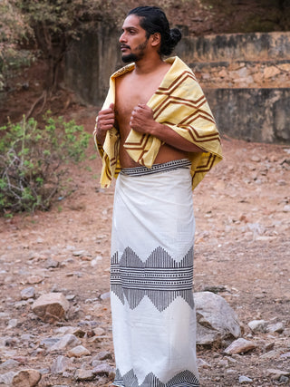 A man stands outdoors on rocky ground, wearing the Arya Dhoti styled traditionally with a block print design and a yellow towel over his shoulders. He looks to the side, with shrubs and a weathered wall behind him.