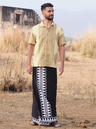 A man stands on dry grassland in a patterned yellow shirt, Jiva Dhoti, and sandals, with an old building and hills in the background.