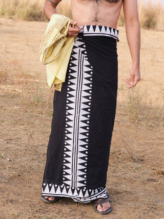 A person wearing a black and white patterned Jiva Dhoti stands outdoors on dry grass, holding a yellow cloth and wearing sandals. The upper body is partially visible with one tattooed arm.