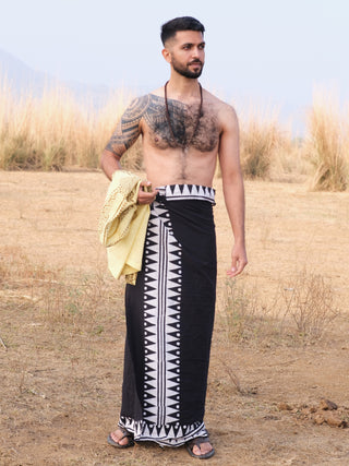 A shirtless man with a right-arm tattoo stands on dry grass, wearing the black and white patterned Jiva Dhoti, holding a yellow cloth and looking to his left.