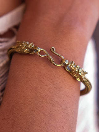 The Takshaka Bracelet, featuring intricately detailed dragon heads at the clasp, graces a medium-toned wrist. This premium accessory showcases a stylish blend of elegance and artistry with its textured design.