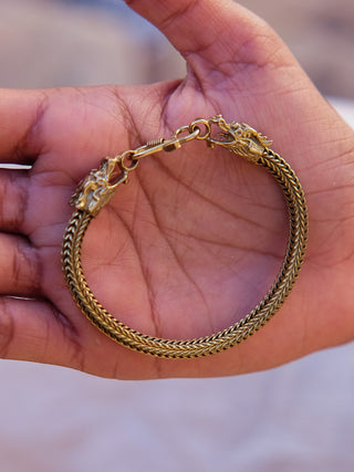 A hand displays the Takshaka Bracelet, a gold-toned accessory with intricately woven design and dragon head clasps. The bracelet gleams brilliantly, reflecting its premium craftsmanship against a blurred background that elegantly highlights it.