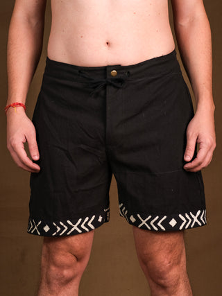 Wearing Tribal Shorts in black with white geometric hem patterns, made from comfortable cotton, the person stands against a brown background with relaxed arms and only their torso and legs visible.