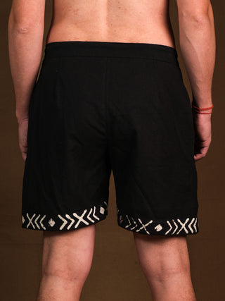 A shirtless person with a red beaded bracelet on their right wrist stands facing away from the camera, wearing black Tribal Shorts adorned with a white Rajasthani hand block print along the bottom hem.