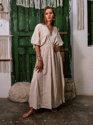 Wearing the Rusalka Maxi Dress in organic khadi cotton, a woman stands barefoot on a rustic floor with green doors and woven wall hangings behind her. Gold jewelry adds to her neutral, relaxed look—perfect effortless boho chic.