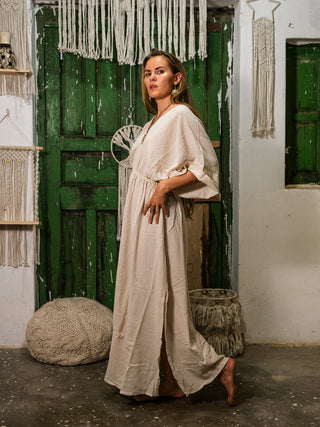 A woman wearing the Rusalka Maxi Dress stands barefoot by a rustic green door, surrounded by woven and macrame decor in a boho chic room.