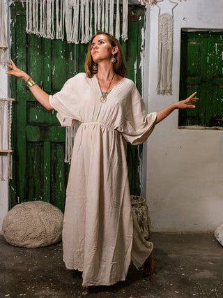 Wearing the Rusalka Maxi Dress, a woman poses gracefully indoors on one foot, arms extended before green rustic doors, surrounded by macramé decor and woven baskets.