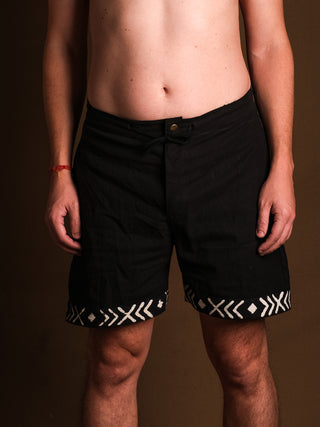 A person stands against a plain brown background, wearing Tribal Shorts featuring white geometric hem patterns on comfortable cotton. The image shows their torso and legs; their face is not visible.