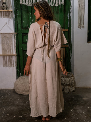 A woman with long hair stands barefoot indoors, facing away, wearing the Rusalka Maxi Dress. The scene features green rustic doors and macramé wall decor in the background.