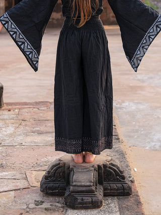 A person models Medusa Palazzo Pants in black with a matching patterned-sleeve top, standing barefoot on a stone pedestal outdoors, arms extended gracefully to the sides.