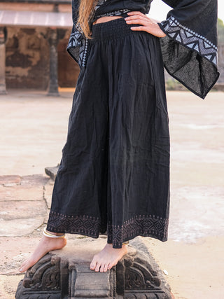 A person in black Medusa Palazzo Pants with Rajasthani handblock print and a matching top poses barefoot on stone, one leg bent, hand on hip, creating a boho-chic vibe.