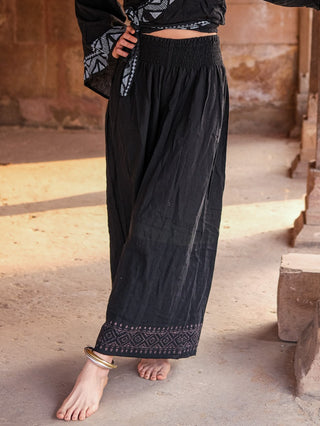 A person wearing Medusa Palazzo Pants with a black top stands barefoot on a stone floor, one foot slightly raised and decorated with a metallic anklet.