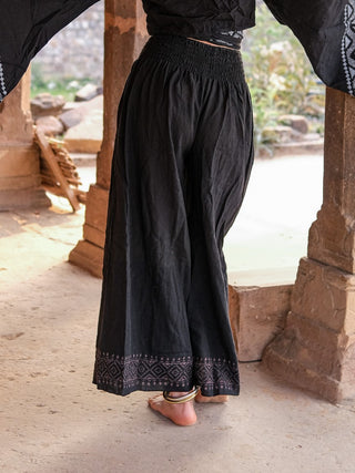 A person stands barefoot on a stone floor, wearing Medusa Palazzo Pants with Rajasthani handblock print hem and a black wide-sleeved top; stone pillars and lush greenery create a serene backdrop.
