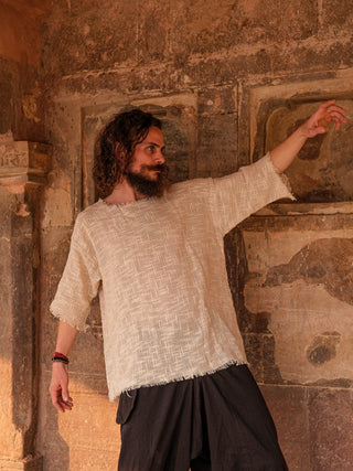 A curly-haired, bearded man poses against a textured stone wall, showcasing his boho style. He wears the Baggy Knit T-shirt with cotton jute fabric and dark pants. His right arm is raised and bent as his left arm extends downward.