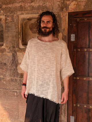 A bearded man with long curly hair stands by an old stone wall in a beige Baggy Knit T-shirt and dark pants, gazing slightly to the side. The wall features rectangular niches and a wooden door with metal details.