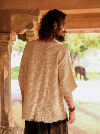 A person with curly hair, wearing a Baggy Knit T-shirt with a boho vibe and arm tattoos, stands under a stone structure, looking away. Sunlight filters through to highlight green foliage and an animal in the background.