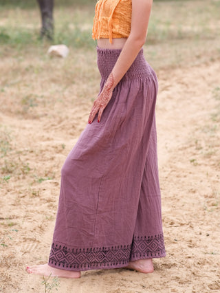 A barefoot person stands sideways on sand, wearing a yellow-orange crop top and Medusa Palazzo Pants with black patterns. Their henna-adorned hand rests on their hip, radiating a boho-chic style.