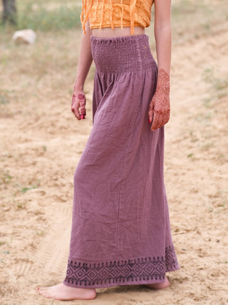 A person stands barefoot on sandy ground, wearing Medusa Palazzo Pants, an orange crop top, and intricate henna designs on their left hand, embodying effortless boho-chic style.