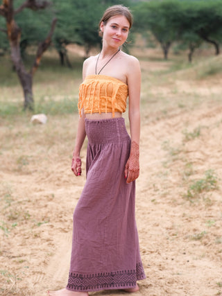 A young woman stands barefoot on a dirt path surrounded by lush greenery, wearing a yellow strapless top and Medusa Palazzo Pants, with intricate mehndi designs adorning her hands.