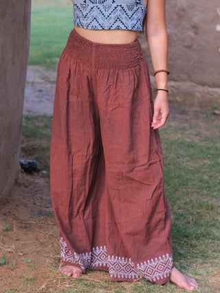 Barefoot on grass, a person models Medusa Palazzo Pants—loose and brown with white hem embroidery—and a blue patterned crop top; their head is out of frame, evoking a relaxed boho-chic style.
