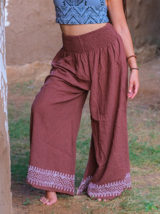 A person poses barefoot outdoors near a mud wall, embracing boho-chic style in brown Medusa Palazzo Pants with white Rajasthani handblock print at the hems, paired with a blue patterned crop top and a beaded bracelet.