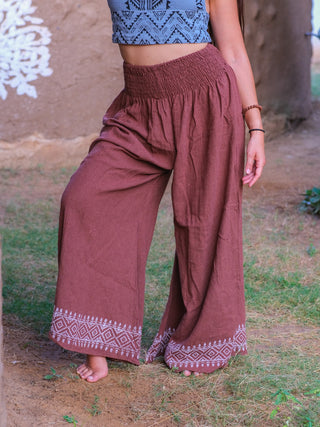 A woman stands barefoot outdoors, wearing Medusa Palazzo Pants with white geometric embroidery at the hem and a blue patterned crop top. Her face is not visible.