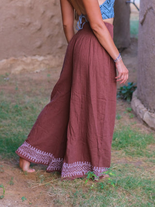 A woman stands barefoot on grass near a stone wall, wearing Medusa Palazzo Pants with white geometric hem patterns and a blue crop top. Her face is hidden, spotlighting the boho-chic Rajasthani handblock print style of the pants.