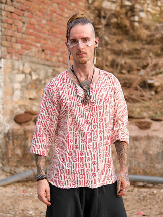 A person with tattoos and facial piercings stands outdoors in a hand block printed red and white Moche Kurta made from 100% cotton, paired with black pants, styled hair, looking at the camera with a brick wall and dry plants in the background.