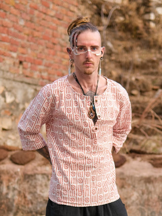 A person with light skin, dreadlocks, facial piercings, and beaded hair stands outdoors wearing glasses and a 100% cotton Moche Kurta. They accessorize with necklaces and look directly at the camera against a blurred brick wall.