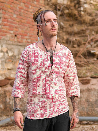 A man with dreadlocks, facial piercings, glasses, and jewelry wears the Moche Kurta—100% cotton hand block printed in a red and white pattern—while standing outdoors by a brick and stone wall, looking slightly upward.