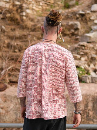 A person with dreadlocks and beaded hair accessories, seen from behind outdoors amid dry foliage and rocks, wears the Moche Kurta—a loose, hand block printed 100% cotton piece with red and white geometric designs—paired with black pants.
