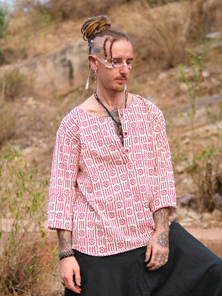 A person with dreadlocks, facial jewelry, and tattoos wears a hand block printed red and white Moche Kurta made from 100% cotton with black pants, standing outdoors in a dry, grassy area with rocks behind them.
