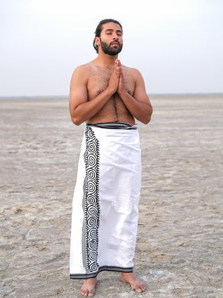 A man stands barefoot on a flat, arid landscape with his hands in prayer. He wears a Tribal Dhoti featuring Rajasthani hand block-print patterns wrapped around his waist, leaving his upper body uncovered. The background is a broad expanse of dry land under a clear sky.