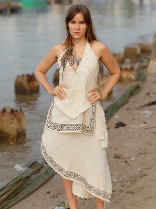 A woman with long brown hair stands on a sandy beach near the water, hands on hips. She wears the Triveni Top by HWC, a beige asymmetrical top featuring intricate block print patterns and a subtle v-neck design. Wooden posts can be seen in the background.