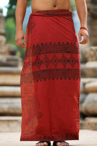 Wearing the Vasuki dhoti, a person is draped in red cotton with intricate black geometrics and florals from Rajasthani handblock printing. They stand in an outdoor setting with their arms and feet visible against an ancient stone structure.