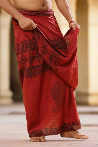 A person stands barefoot in a Vasuki dhoti, adorned with black Rajasthani handblock prints, gently holding the fabric at the waist. The softly blurred background hints at a warm, stone architectural setting, evoking crystal healing serenity.