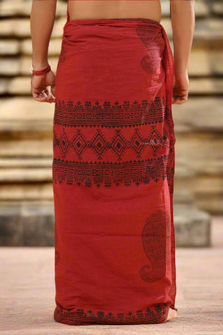 Wearing the Vasuki dhoti, a person stands on stone steps, showcasing its red fabric adorned with black geometric and paisley patterns reminiscent of Rajasthani handblock print. The image highlights the back view of this stunning attire.