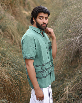 A man with dark hair and a beard stands in tall grass, wearing the green Deva Shirt featuring a geometric pattern and paired with white pants. He looks at the camera, lightly touching his collar.