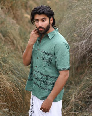 A man with dark hair and a beard stands in tall grass, wearing the premium cotton Deva Shirt in green with black geometric patterns and a white garment. He poses with his hand near his face, highlighting the shirts unique style.