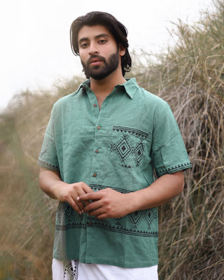 A man with medium-length dark hair and a beard stands outdoors in tall grass, wearing the green Deva Shirt—crafted from premium cotton with geometric block print—and white pants. He gazes calmly at the camera.