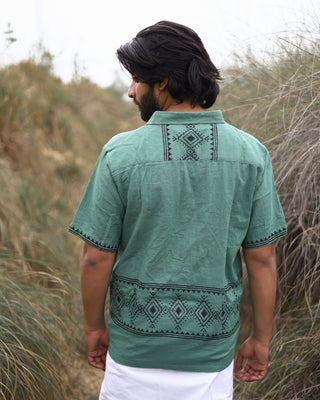 A man with medium-length dark hair stands among tall grasses, seen from behind. He’s wearing the Deva Shirt—a green shirt with black geometric patterns—and white pants.
