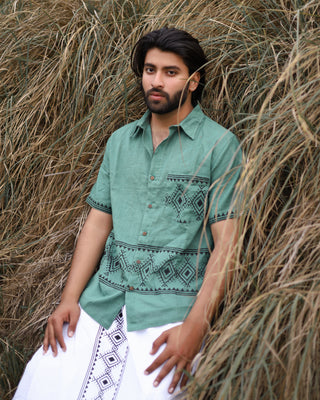 A man with medium-length dark hair and a beard stands against tall, dry grass, wearing the Deva Shirt—green with geometric block prints—and a white garment with black designs draped around his waist.