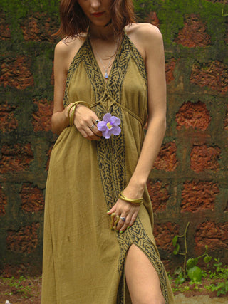 Wearing the green Devi Maxi Dress, this person stands by a red stone wall holding a purple flower. The sleeveless patterned dress is crafted from organic cotton, and their partially visible face is adorned with rings and bangles.