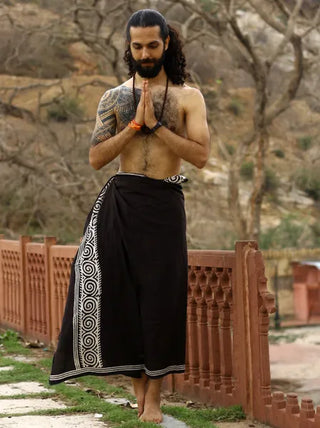 A bearded, tattooed person stands barefoot on grass, wearing a black Tribal Dhoti styled from a Rajasthani hand block-print wrap. With hands in prayer position, bare trees and a wooden railing frame the serene outdoor scene.