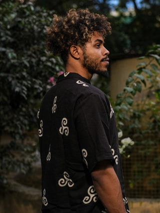 A man with curly hair and a beard stands in profile wearing a Spiral Kurta, surrounded by greenery in a dimly lit outdoor setting.